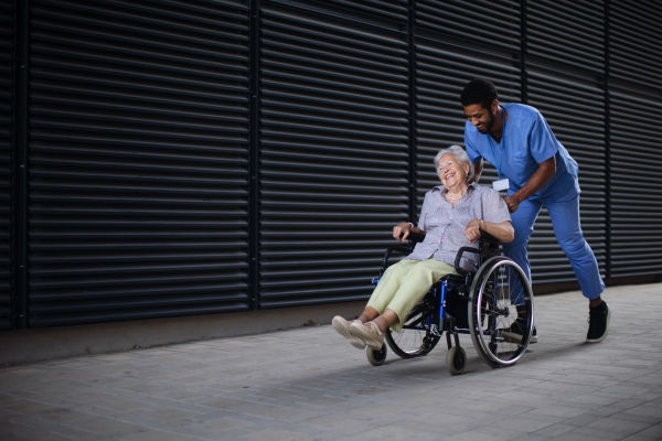 Caregiver man pushing excited senior woman at wheelchair on a street, enjoying time together.