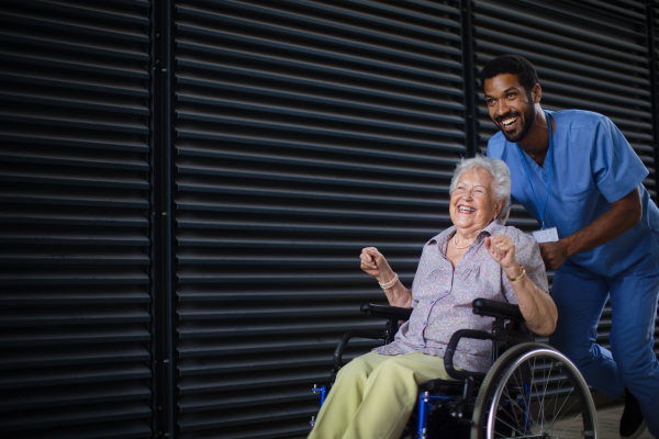 Caregiver man pushing excited senior woman at wheelchair on a street, enjoying time together.