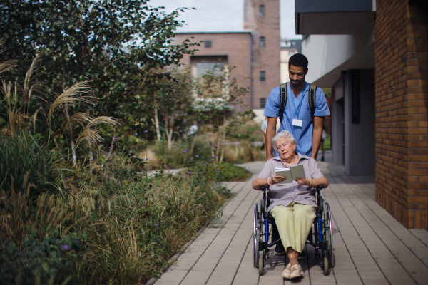 Caregiver man pushing senior woman at a wheelchair on the street.