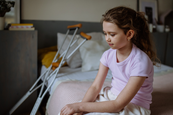 Little unhappy girl with broken leg sitting on a bed in her room.