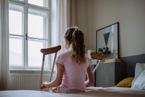 Rear view of little girl with broken leg sitting on a bed in her room.