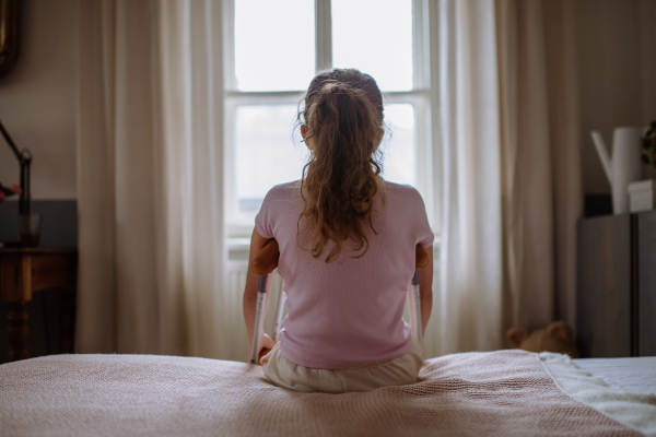 Rear view of little unhappy girl with broken leg sitting on a bed in her room.