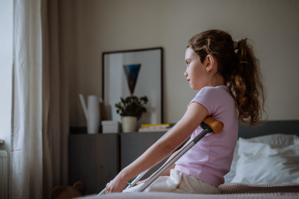 Little unhappy girl with broken leg sitting on a bed in her room.