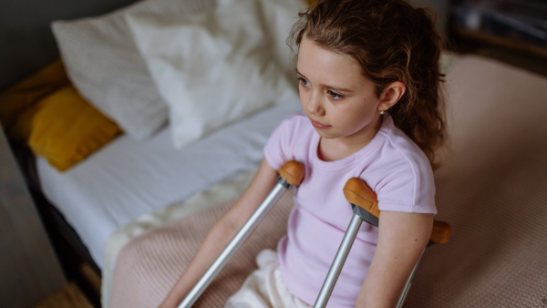 Little unhappy girl with broken leg sitting on a bed in her room.