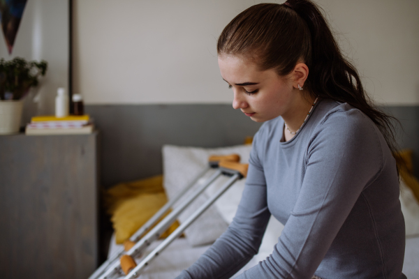 Unhappy teenage girl with broken leg sitting on a bed in her room.