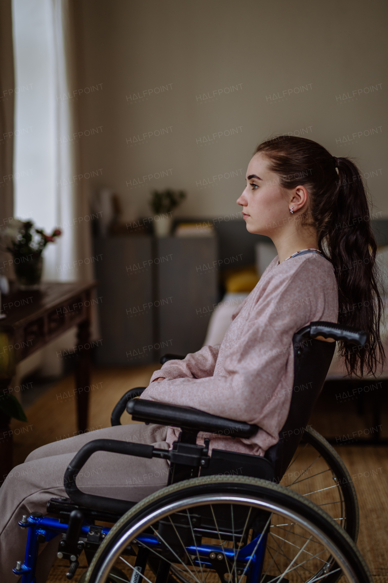 Unhappy teenage girl sitting on wheelchair and looking out of the window.