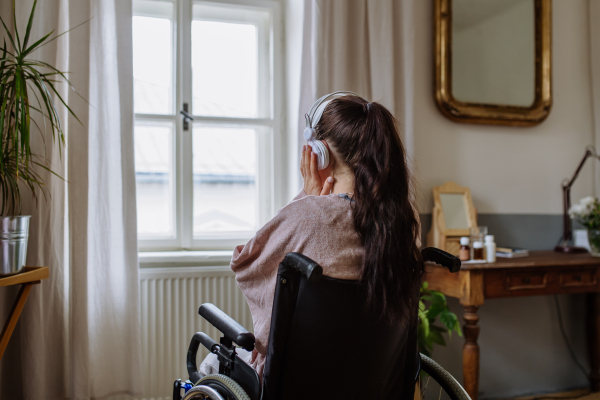 Teenage girl sitting on wheelchair, listening music and looking out of the window.