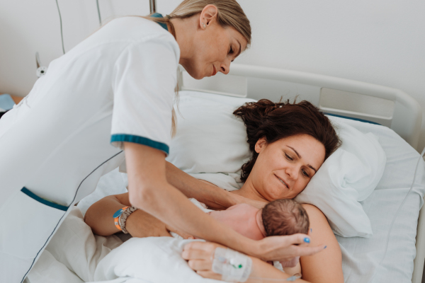 Lactation consultant helping with breastfeeding newborn baby, latching, feeding on first milk, colostrum. Happy mother bonding with newborn baby right after birth. Skin to skin contact.