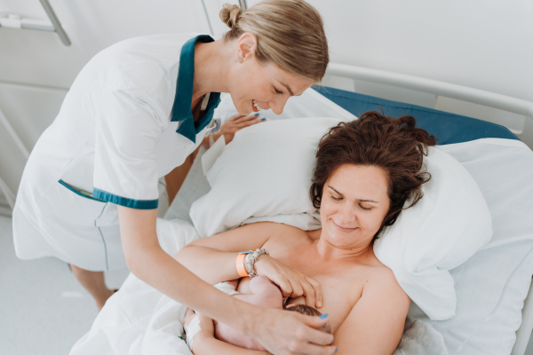 Lactation consultant helping with breastfeeding newborn baby, latching, feeding on first milk, colostrum. Happy mother bonding with newborn baby right after birth. Skin to skin contact.