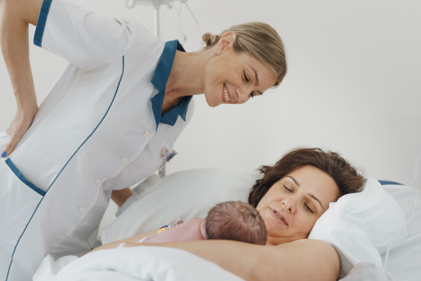 Lactation consultant helping with breastfeeding newborn baby, latching, feeding on first milk, colostrum. Happy mother bonding with newborn baby right after birth. Skin to skin contact.