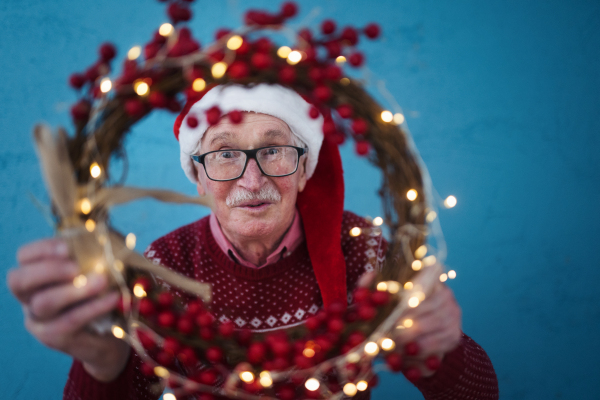 Portrait of happy senior man with Santa Claus hats in studio shoot, looking trough Christmas wreath.