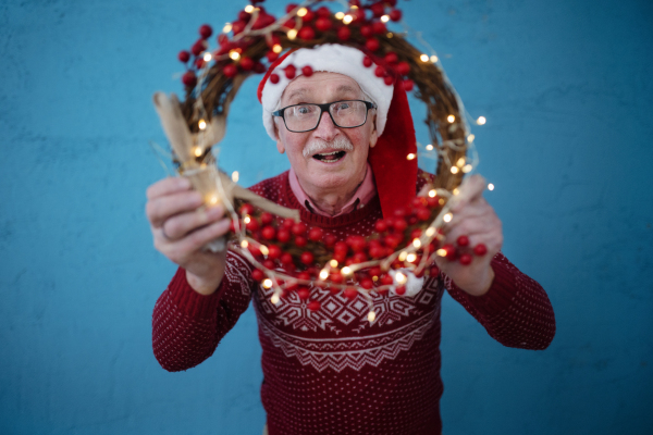 Portrait of happy senior man with Santa Claus hats in studio shoot, looking trough Christmas wreath.