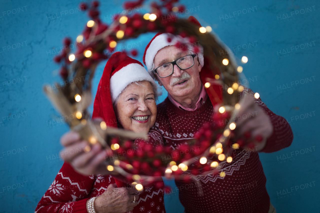 Portrait of happy senior couple with Santa Claus hats in studio shoot, looking trough Christmas wreath.