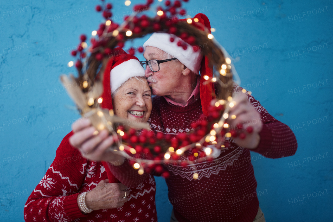 Portrait of happy senior couple with Santa Claus hats in studio shoot, looking trough Christmas wreath.