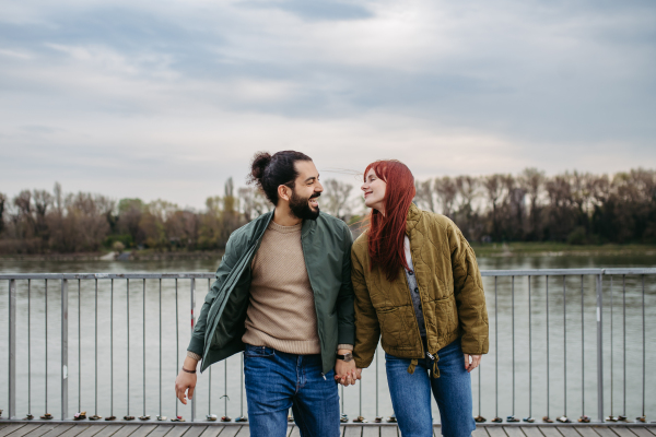 Beautiful couple on romantic date in the city, walking along the boardwalk by the river, holding hands. Together during cold spring, autumn day.