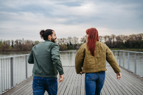 Couple on romantic date in the city, walking along the boardwalk by the river. Rear view. Together during cold spring, autumn day.