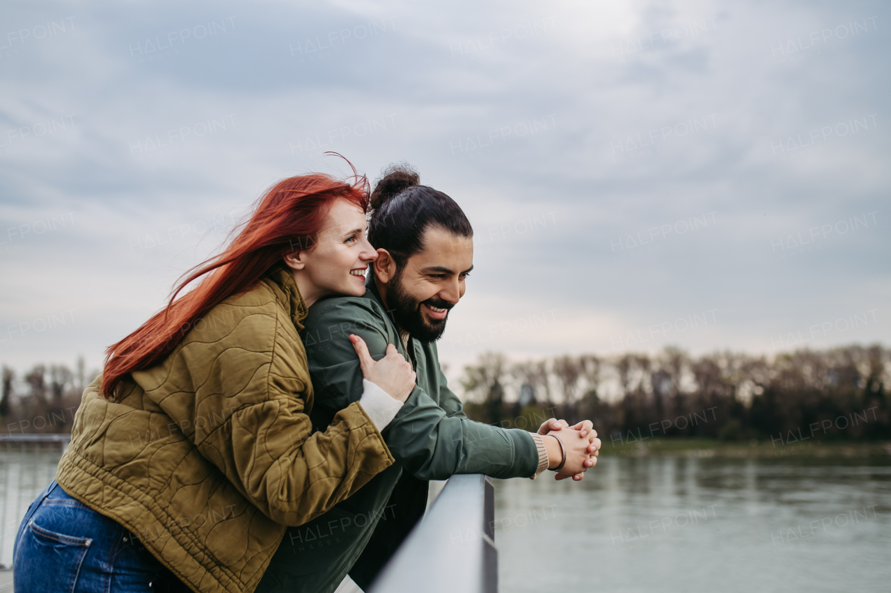 Beautiful couple on romantic date in the city, standing on boardwalk by the river, looking at water. Together during cold spring or autumn day.