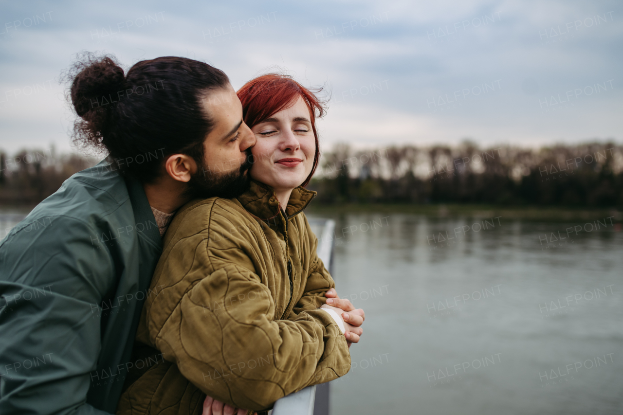 Beautiful couple on romantic date in the city, standing on boardwalk by the river, looking at water. Kissing on cheek