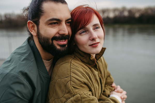 Beautiful couple on romantic date in the city, standing on boardwalk by the river, looking at water. Together during cold spring or autumn day.