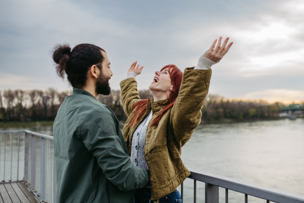 Beautiful couple on romantic date in the city, standing on boardwalk by the river. Hands up in the air.