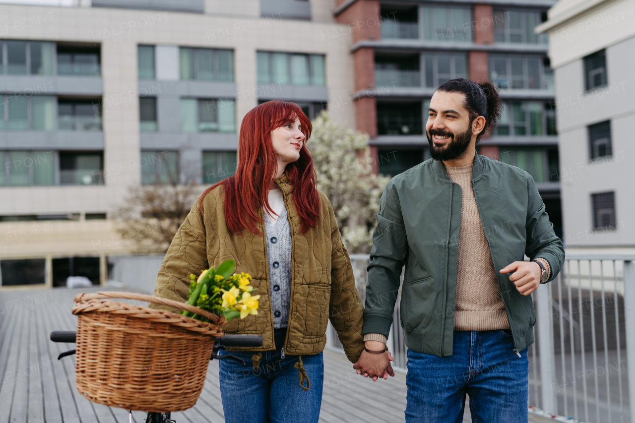 Beautiful couple on walk along the boardwalk by the river, holding hands. Woman pushing bicycle with basket. Together on romantic date during cold spring or autumn day