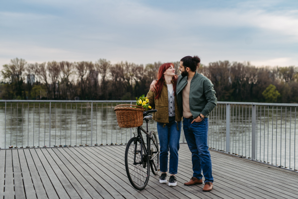 Couple in love on walk along the boardwalk by the river, man with arm around girlfiriend shoulders. Woman pushing bicycle with basket.