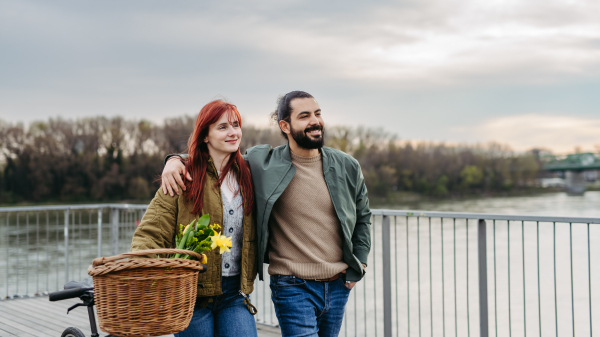 Couple in love on walk along the boardwalk by the river, man with arm around girlfiriend shoulders. Woman pushing bicycle with basket.