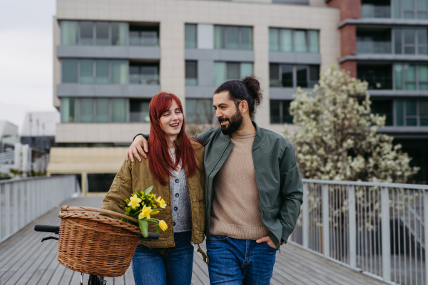 Couple in love on walk along the boardwalk by the river, man with arm around girlfiriend shoulders. Woman pushing bicycle with basket.