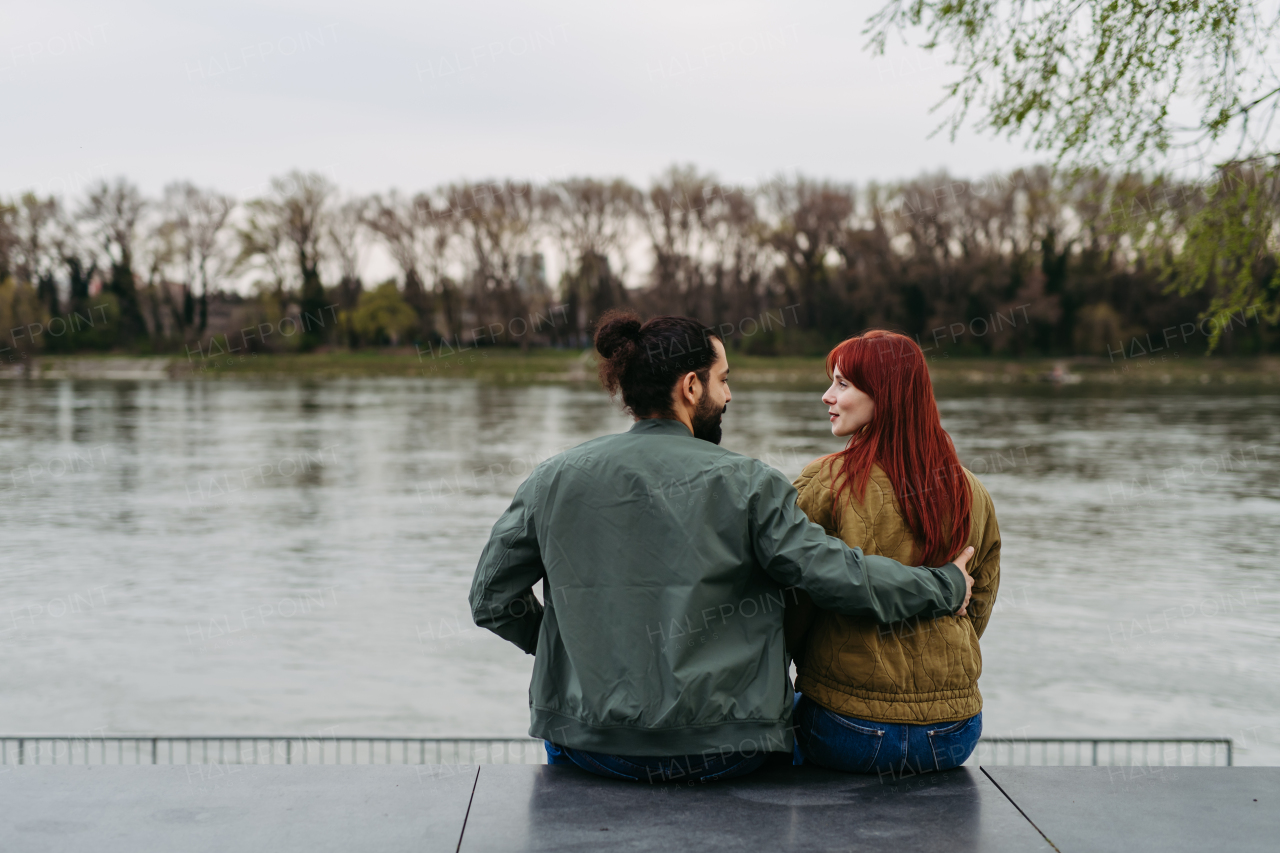 Couple on romantic date in the city, sitting on riverbank and looking at each other. Rear view. Together during cold spring or autumn day by river