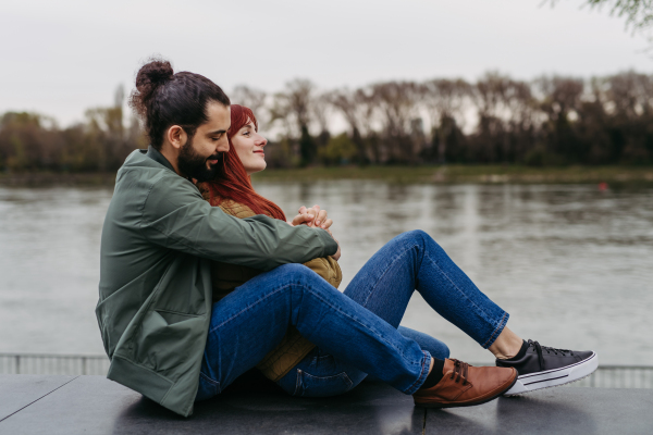 Couple on romantic date in the city, sitting on riverbank. Man hugging beautiful girlfriend. Together during cold spring or autumn day by river