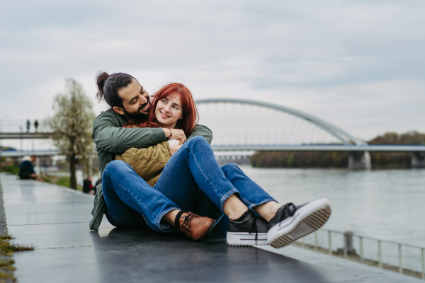 Couple on romantic date in the city, sitting on riverbank. Man hugging beautiful girlfriend. Together during cold spring or autumn day by river