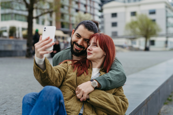 Couple on romantic date in the city, sitting on riverwalk, taking selfie. Man hugging beautiful girlfriend. Together during cold spring or autumn day by river