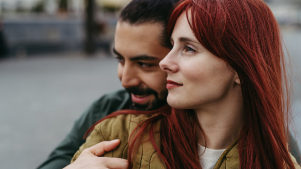 Couple on romantic date in the city, sitting on riverbank. Man hugging beautiful girlfriend. Together during cold spring or autumn day by river