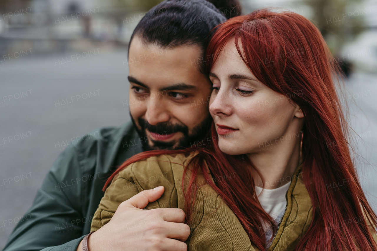 Couple on romantic date in the city, sitting on riverbank. Man hugging beautiful girlfriend. Together during cold spring or autumn day by river