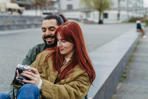 Couple on romantic date in the city, sitting on riverwalk, looking at photos in camera. Together during cold spring or autumn day by river