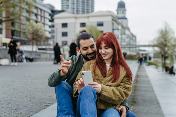 Couple on romantic date in the city, sitting on riverwalk, watching video on smartphone. Together during cold spring or autumn day by river