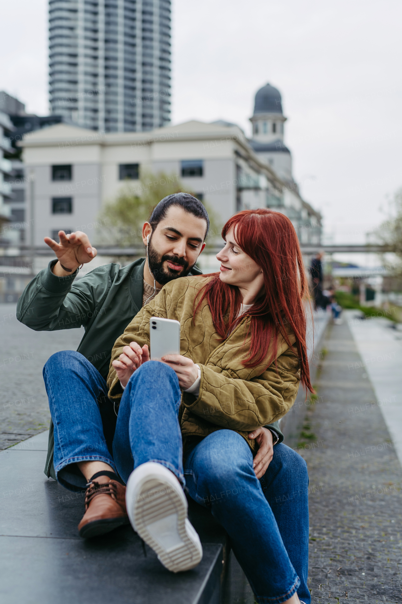 Couple on romantic date in the city, sitting on riverwalk, watching video on smartphone. Together during cold spring or autumn day by river