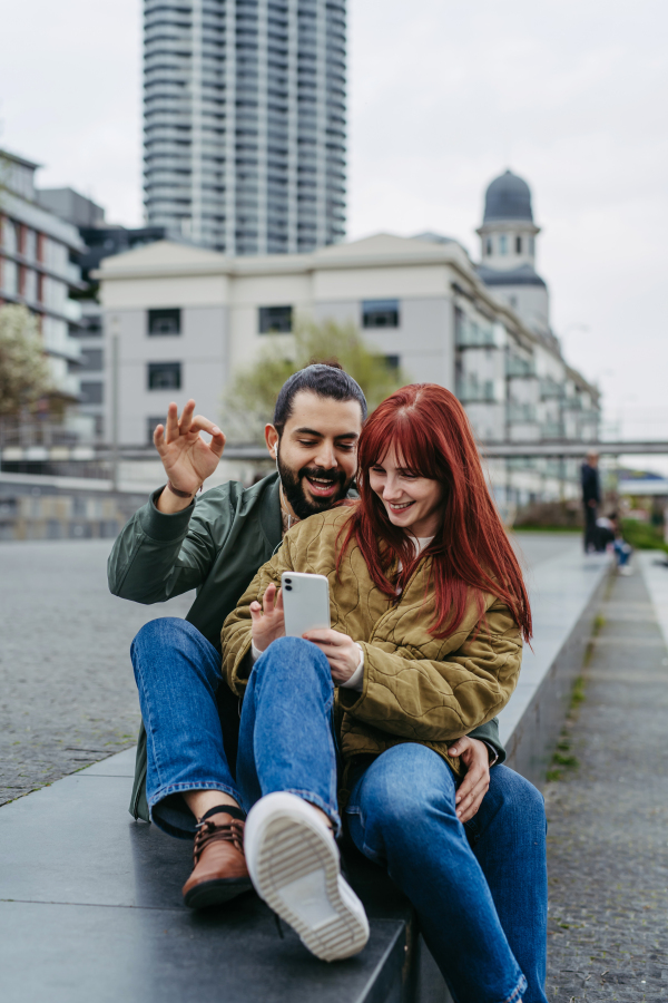 Couple on romantic date in the city, sitting on riverwalk, watching video on smartphone. Together during cold spring or autumn day by river