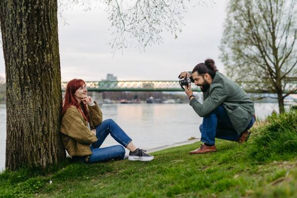 Photographer taking picture of a beautiful model. Woman is sitting on grass by a tree, man is photographing her.