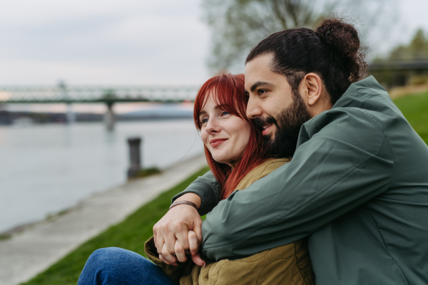 Couple on romantic date in the city, sitting on riverbank. Man hugging beautiful girlfriend. Together during cold spring or autumn day by river