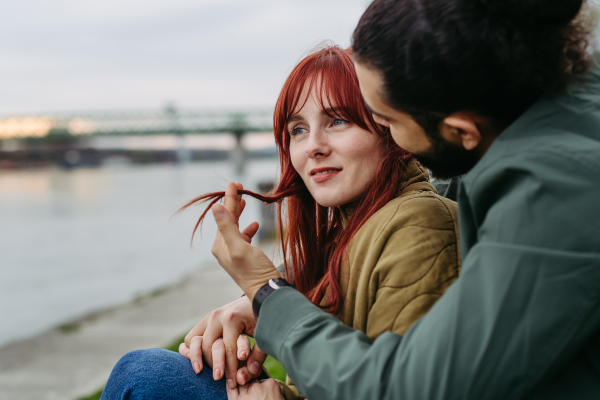 Couple on romantic date in the city, sitting on riverbank. Man playing with girlfriend's hair. Together during cold spring or autumn day by river