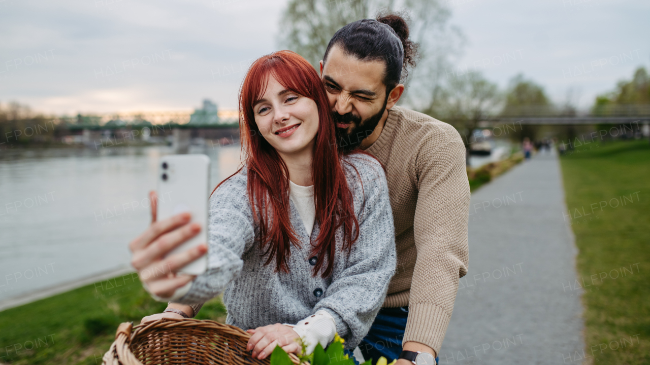 Boyfriend taking selfie with his girlfriend, standing on river embankment. Beautiful couple on romantic date during cold spring or autumn day.