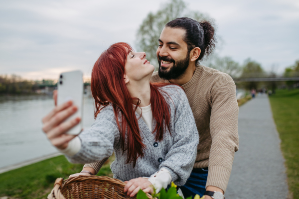 Boyfriend taking selfie with his girlfriend, standing on river embankment. Beautiful couple on romantic date during cold spring or autumn day.