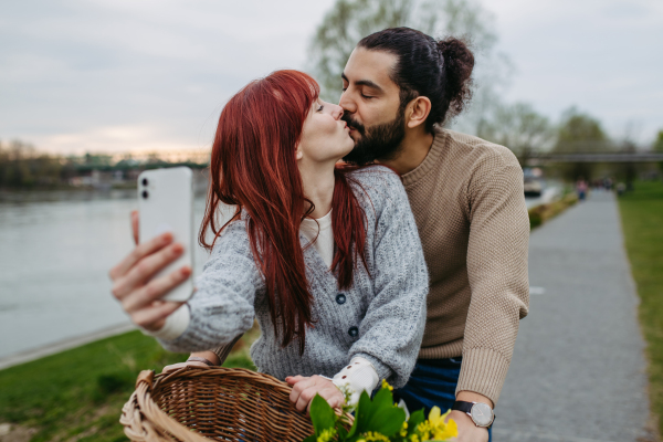 Boyfriend taking selfie with his girlfriend, standing on river embankment. Beautiful couple on romantic date during cold spring or autumn day.