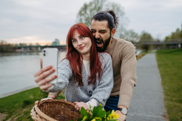 Boyfriend taking selfie with his girlfriend, standing on river embankment. Beautiful couple on romantic date during cold spring or autumn day.