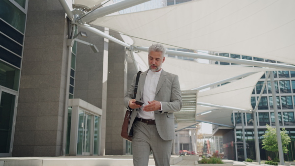 Mature businessman walking outside office building using smart phone in city