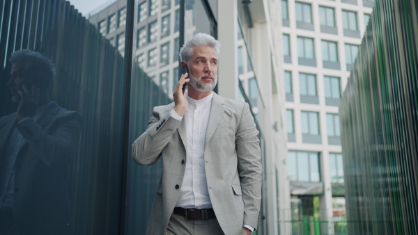 Mature businessman walking outside office building talking on the phone, serious.