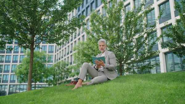 Mature businessman sitting on the grass with book, in front of office building.