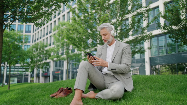 Mature businessman sitting barefoot on grass outside office building, using phone and wearing headphones.