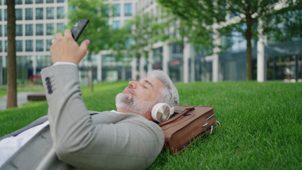 Mature businessman lying on grass outside office building, using phone and wearing headphones, relaxing. Close up.